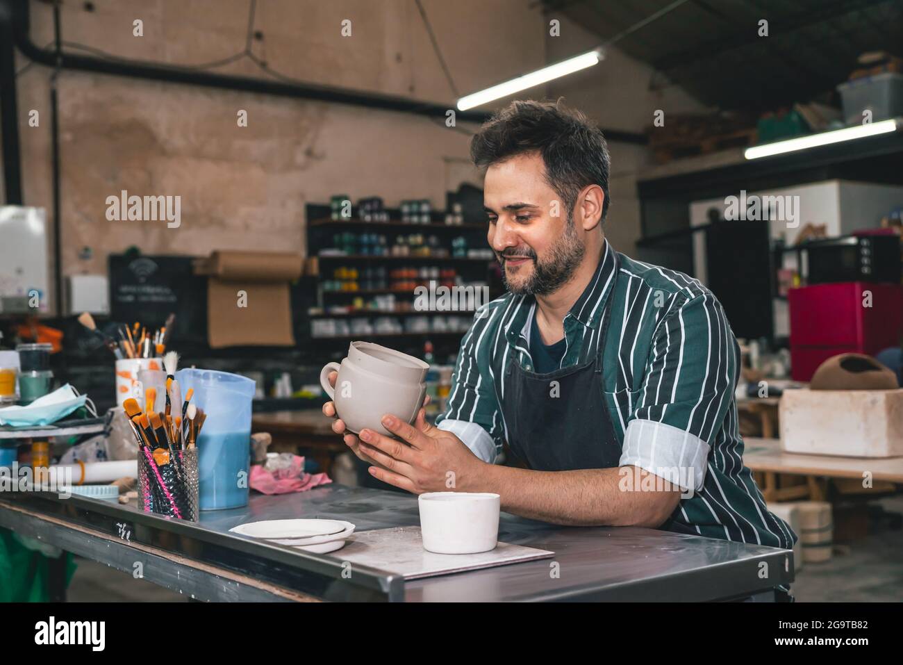 Ein erwachsener hispanischer Mann hält in einer Werkstatt einen Tontopf über einem Tisch Stockfoto