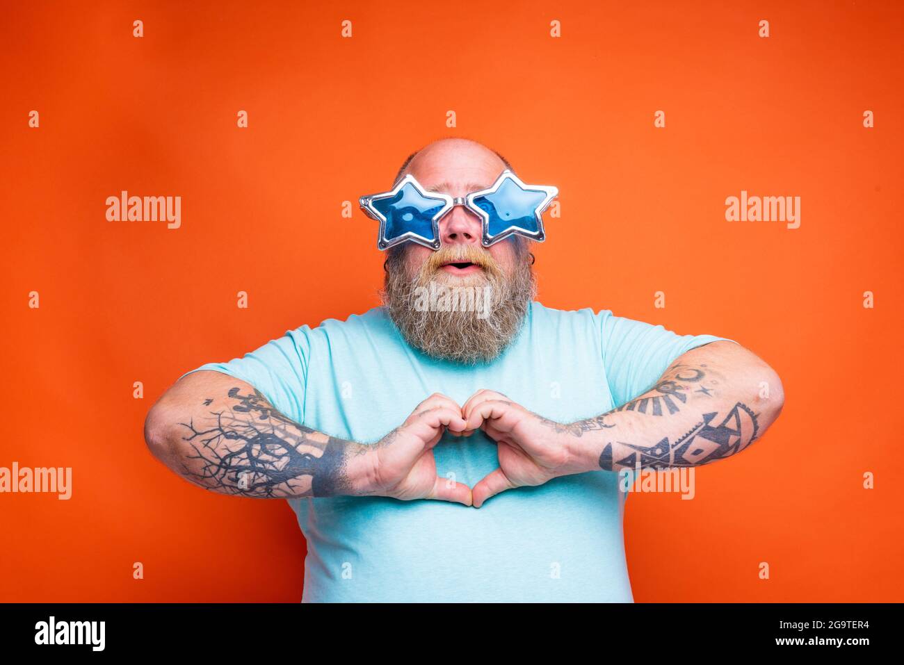 Fetter Mann mit Bart, Tattoos und Sonnenbrille macht mit den Händen Herzform Stockfoto