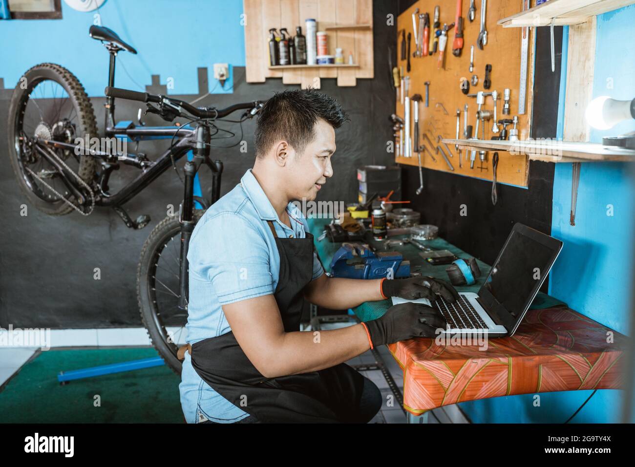 Ein Fahrradmechaniker in einer Schürze, der Handschuhe trägt und einen Laptop benutzt, um nach Ersatzteilen zu suchen Stockfoto