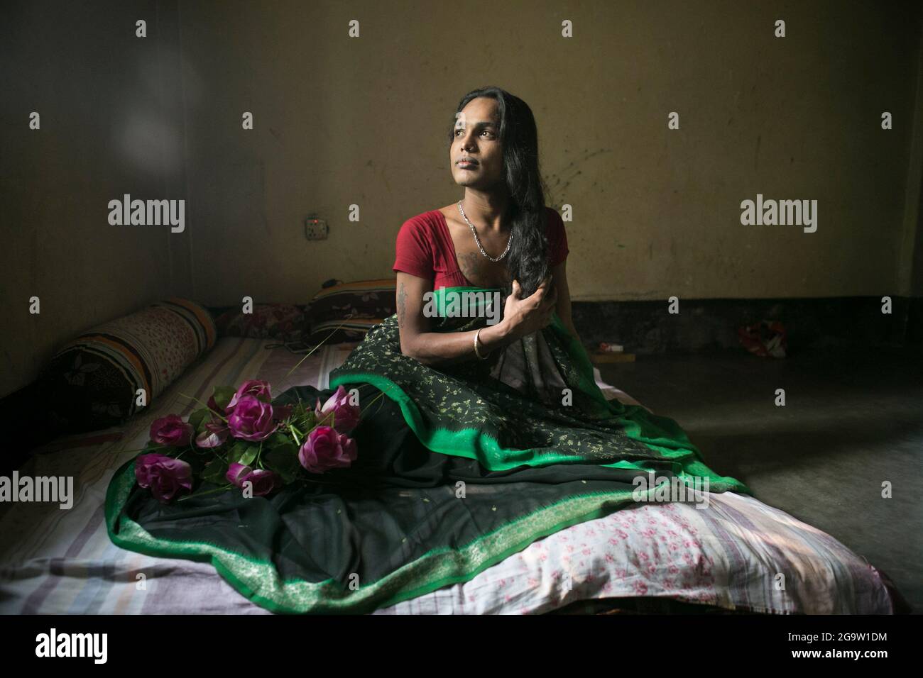 Porträt einer nachdenklichen indischen Transgender-Frau, die drinnen in traditionellem Saree sitzt und Identität und Schönheit in einem ruhigen, schwach beleuchteten Raum vereint. Stockfoto