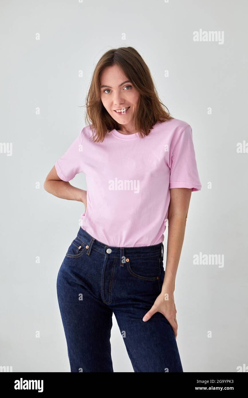 Positive weibliche Modell trägt rosa T-Shirt und Jeans stehen mit der Hand auf der Taille vor weißem Hintergrund und Blick auf die Kamera Stockfoto