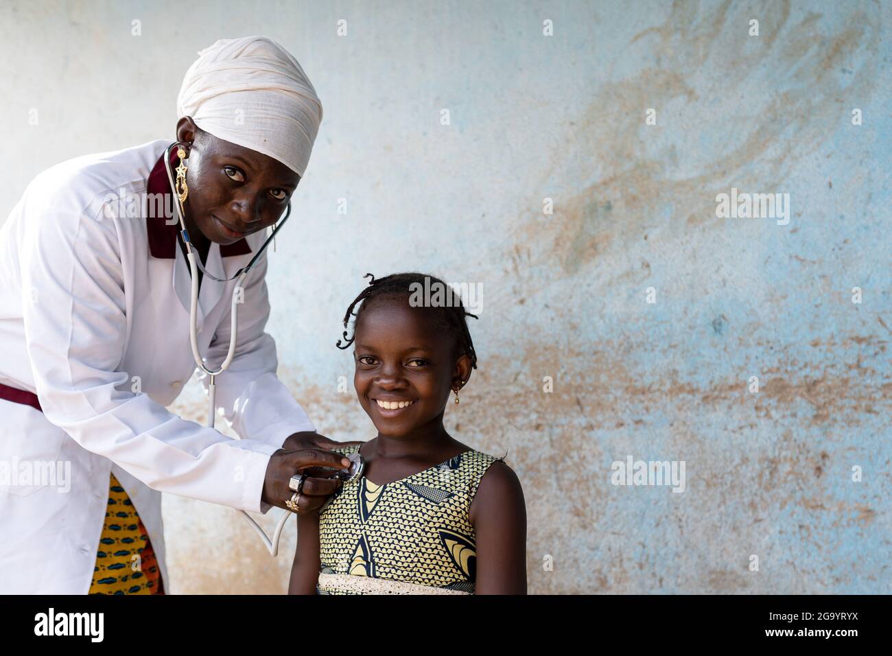 In diesem Bild untersucht eine schwarze Krankenschwester die Lunge und das Herz eines lächelnden, selbstbewussten kleinen afrikanischen Schulmädchen mit einem Stethoskop in einem ländlichen Krankenhaus Stockfoto