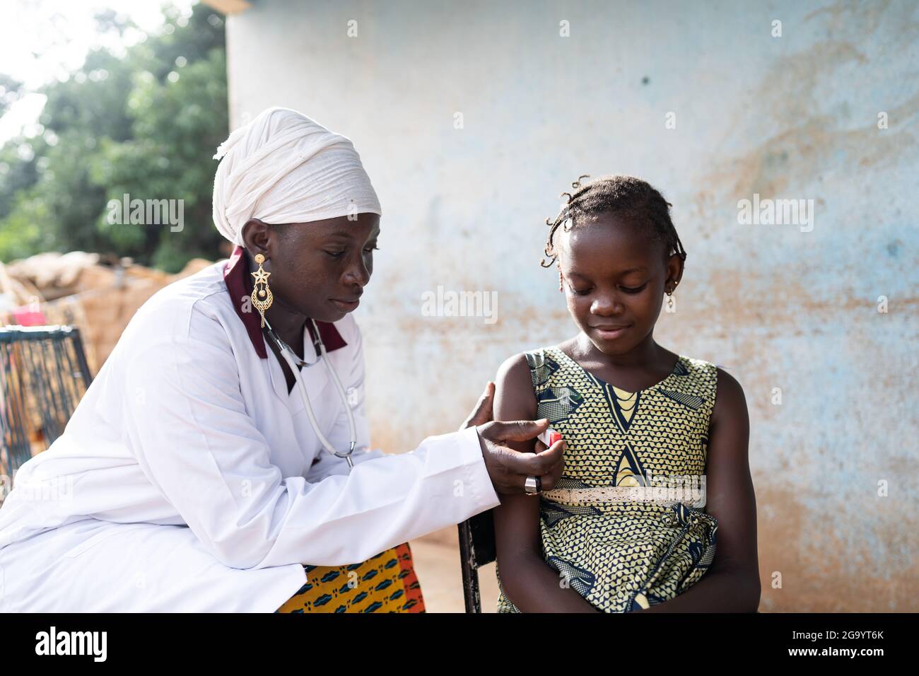 Auf diesem Bild platziert eine aufmerksame und sanfte junge afrikanische Krankenschwester, die in einem weißen Mantel gekleidet ist, ein Digitalthermometer unter die Achsel eines kleinen Schwarzen Stockfoto