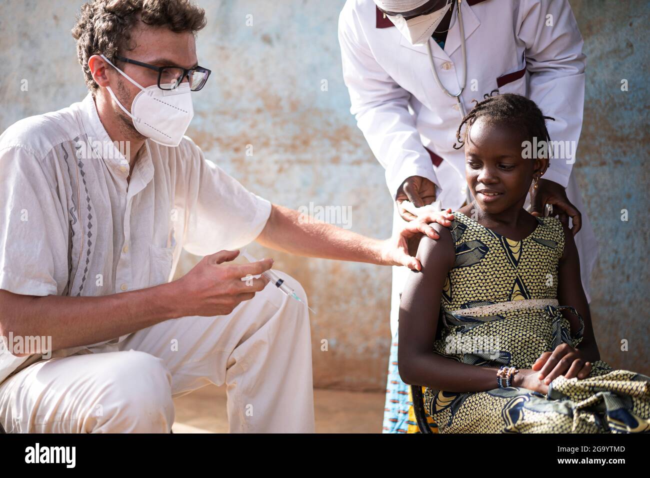 In diesem Bild hält ein sitzender weißer Freiwilliger-Arzt mit Gesichtsmaske eine Spritze in seiner rechten Hand, während er die Haut eines süßen schwarzen sch zentiert Stockfoto