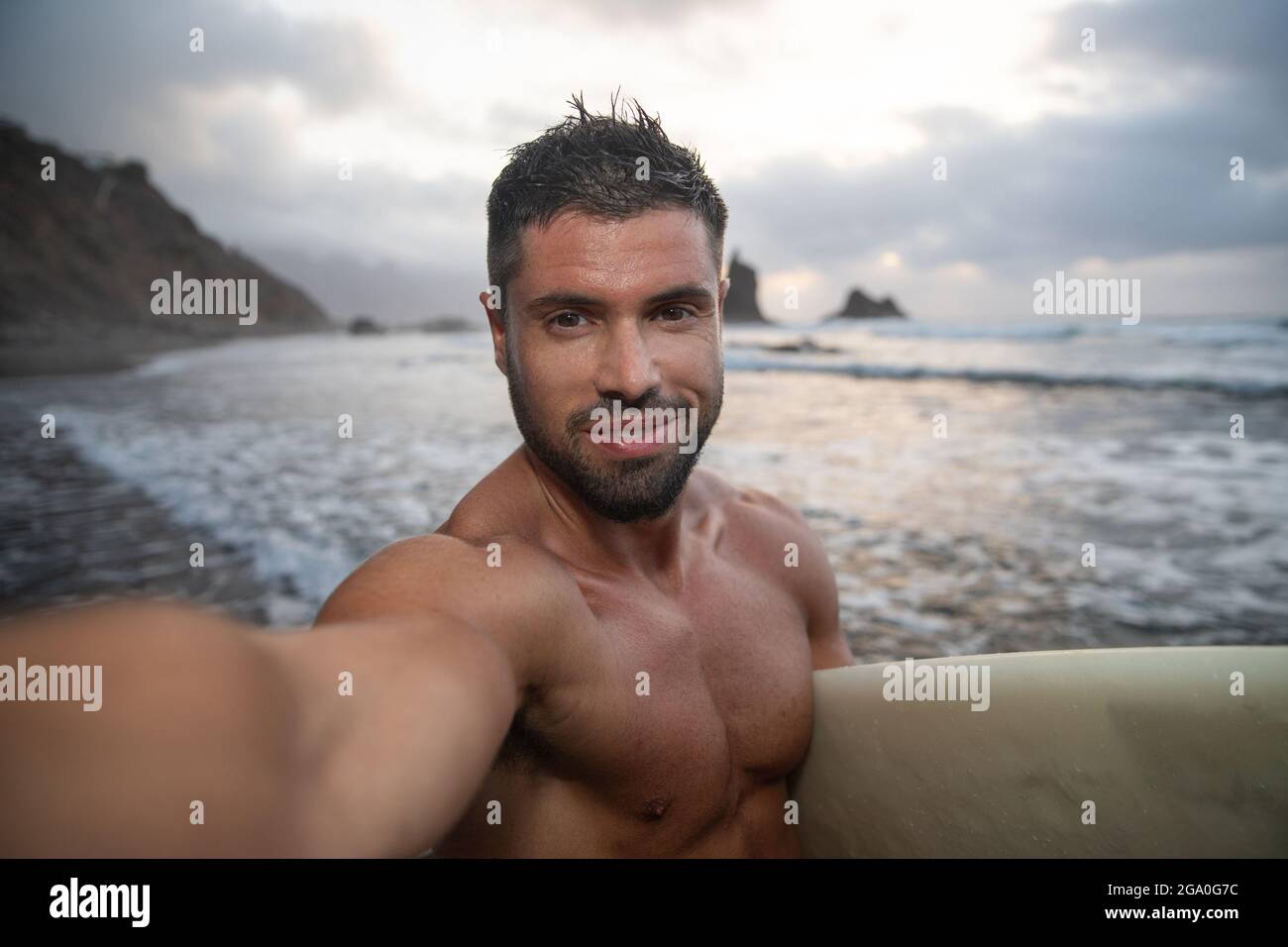 Surfer macht ein Selfie am Strand während des Sonnenuntergangs, er hält sein Surfbrett und lächelt. Junger attraktiver Mann am Strand - fröhliche Person beim Üben Stockfoto