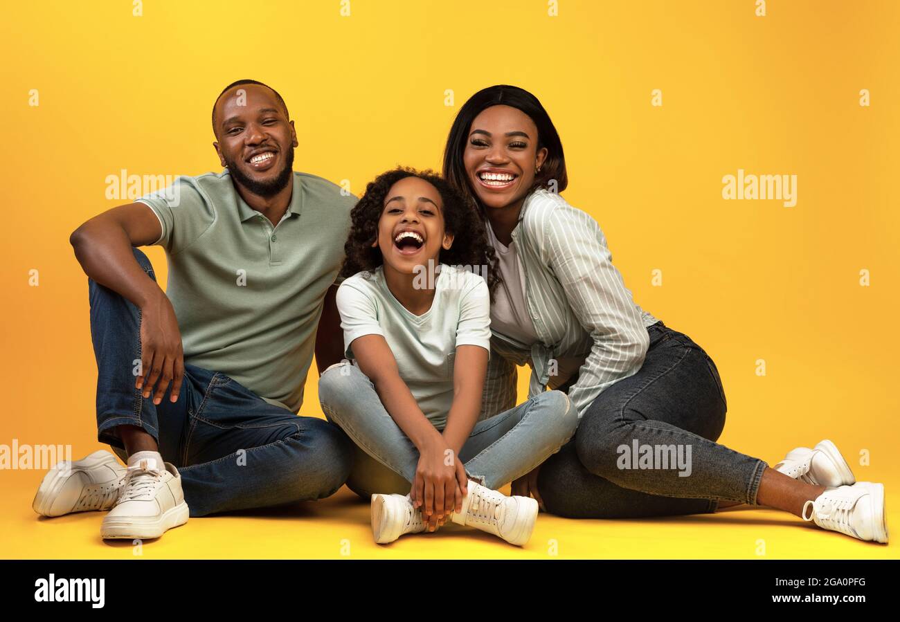 Glückliche afroamerikanische Familie, die auf dem Boden sitzt und lacht, lächelt zusammen vor der Kamera über dem gelben Studiohintergrund Stockfoto