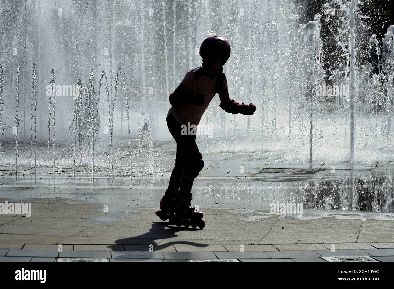 Die Silhouette eines kleinen Mädchens auf dem Rollschuhlaufen kühlt sich im Park unter Wassersprinklern ab, die aufgestellt wurden, um die drückende Sommerhitze in Istanbul, Türkei, zu schlagen Stockfoto