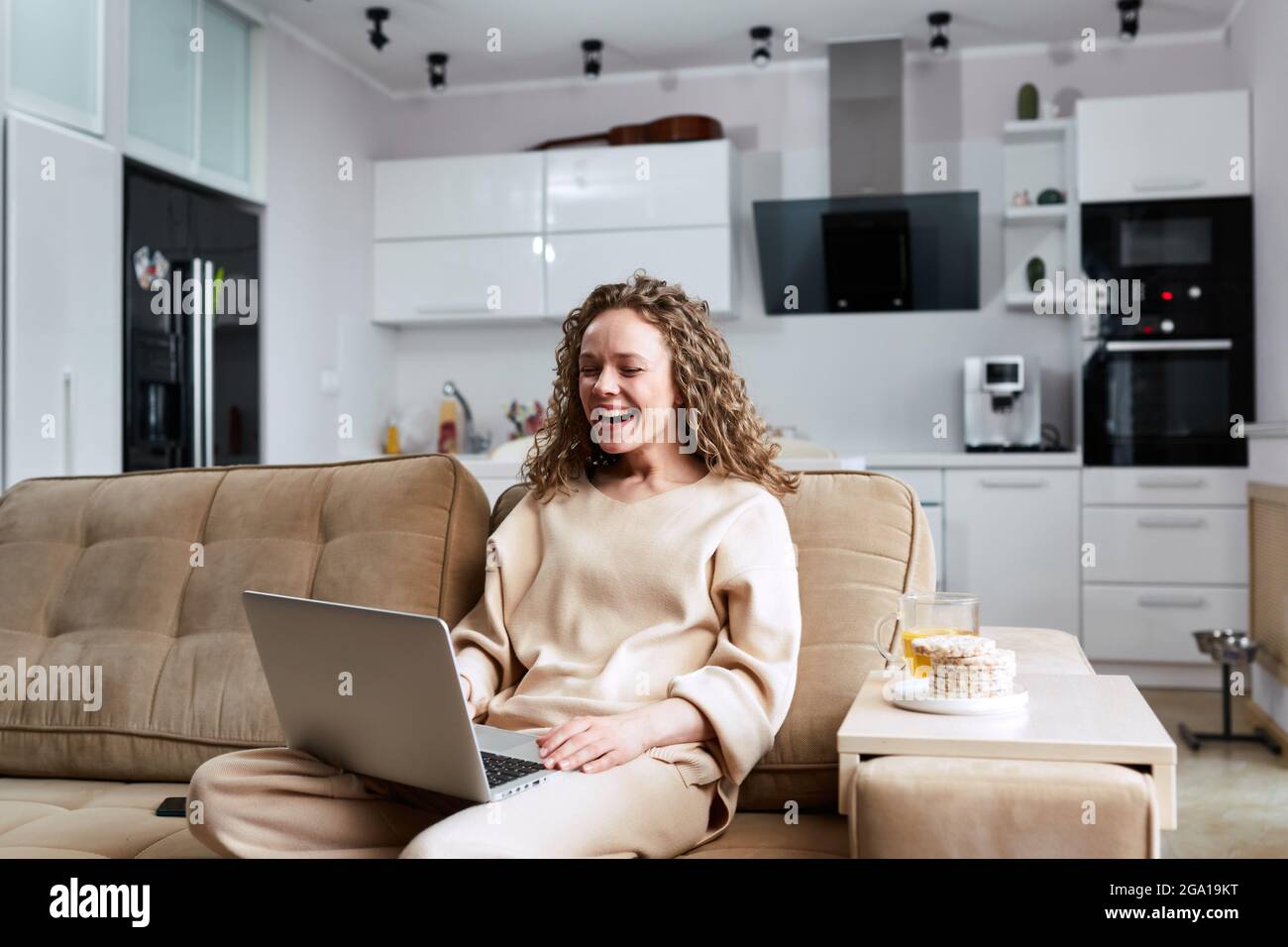 Schöne fröhliche Mädchen mit silbernen Laptop, während auf dem Sofa im Wohnzimmer sitzen. Attraktive Geschäftsfrau, die online studiert, mit Laptop-Software, Web Stockfoto