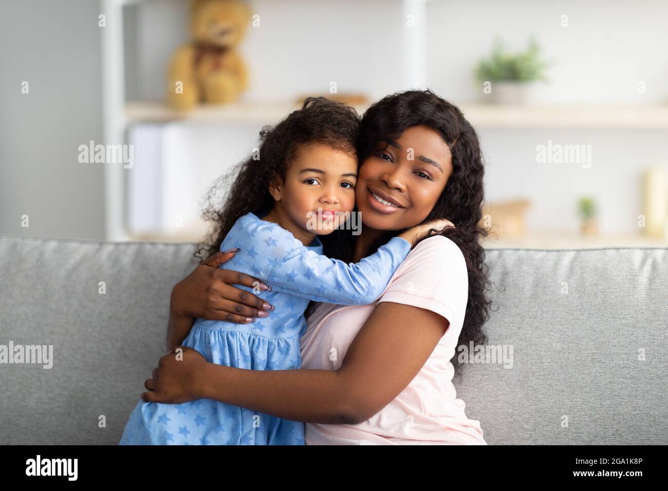 Liebevolle schwarze Mädchen in hübschen Kleid umarmt ihre Mutter auf dem Sofa im Wohnzimmer. Konzept für starke Familienbeziehungen Stockfoto