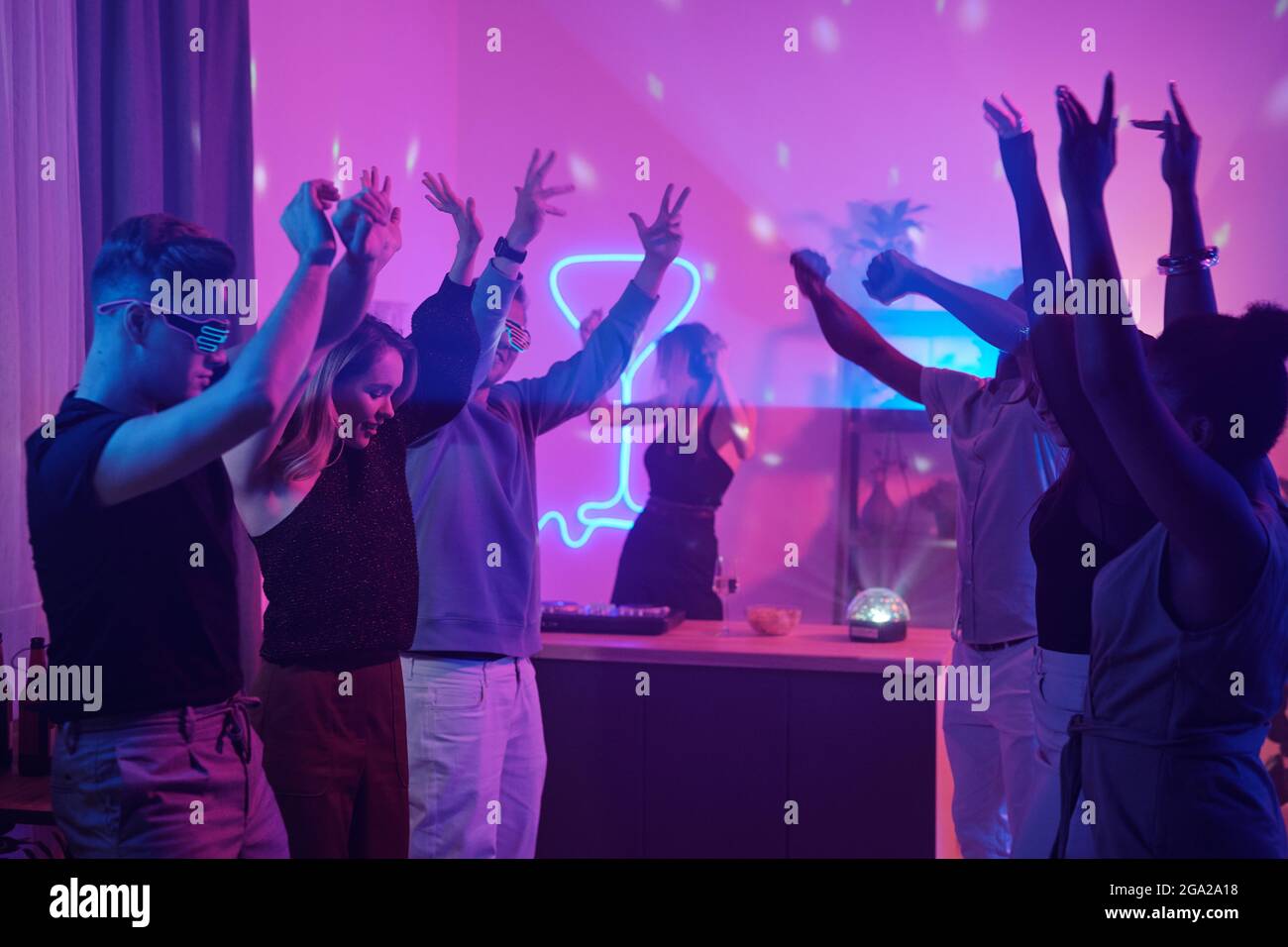 Junge interkulturelle Freunde in smarter Casualwear, die beim Tanzen die Arme heben Gemeinsam zu Hause Party im Wohnzimmer mit rosa Beleuchtung beleuchtet Stockfoto