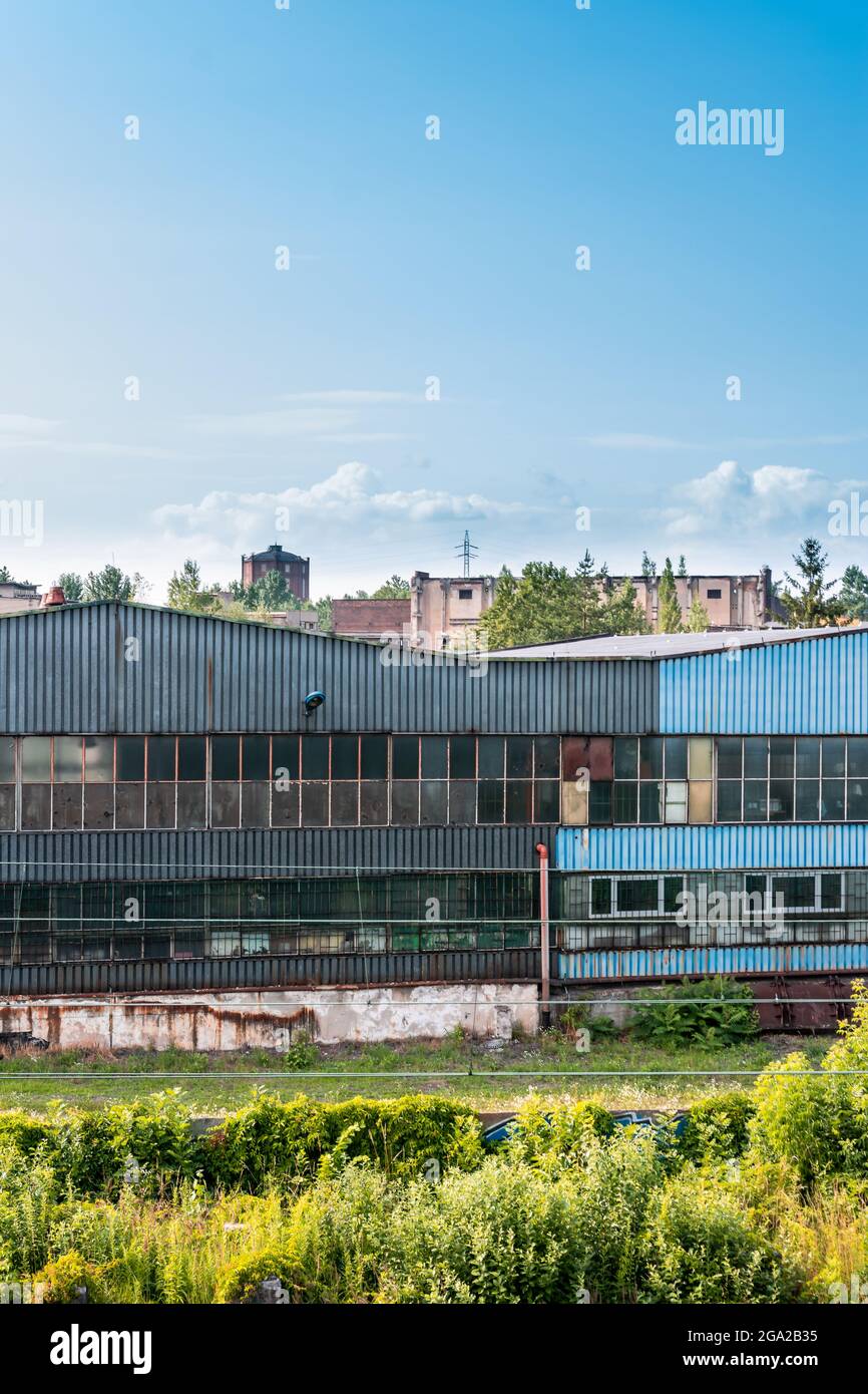 Alte, verrostete Fabrikhalle aus Metall im Industrieviertel einer Stadt. Hohes Gras und Büsche im Vordergrund, Stadtbild und blauer Himmel im Hintergrund Stockfoto