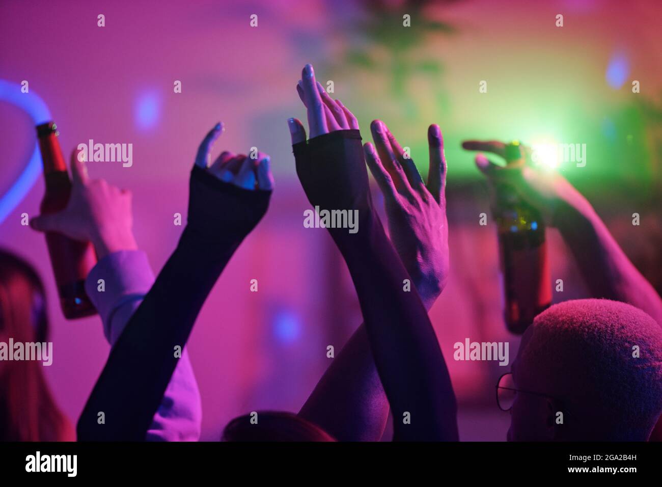 Hochgezogene Arme junger interkultureller Freunde, die zu Hause tanzen Party und Getränke gegen rosa Lichter während des Wochenendes genießen Stockfoto