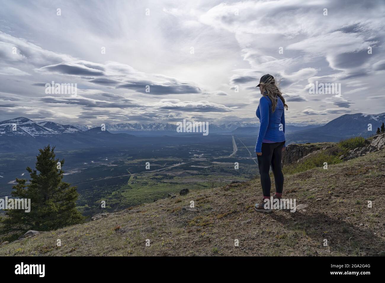 Blick von hinten auf eine Frau, die auf einem Berggipfel steht und auf die aussicht auf die majestätischen Bergketten und das Tal unten mit einem dramatischen... Stockfoto