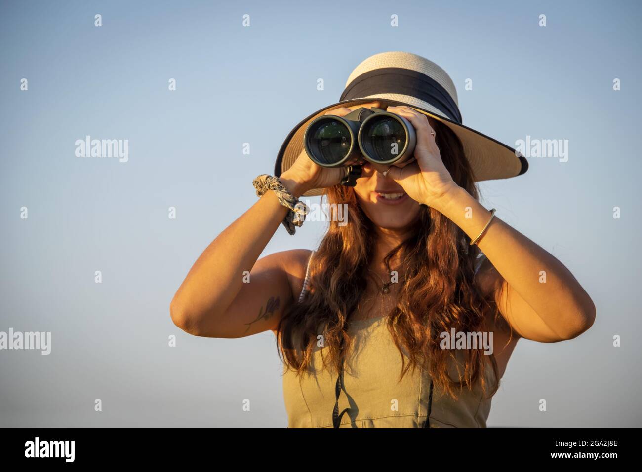 Nahaufnahme einer Frau mit Strohhut und Blick durch ein Fernglas in das helle Sonnenlicht vor einem blauen Himmel auf der Gabus Game Ranch Stockfoto