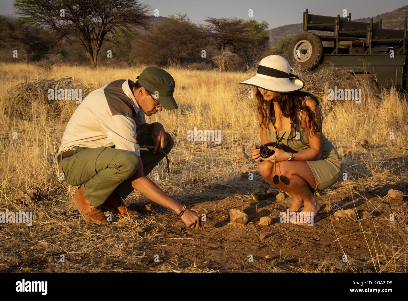 Safariführer mit einer Reisenden Frau, die einen Strohhut trägt und eine Kamera hält, die auf dem Boden hockend auf der Savanne beim Gabus Game Ran... Stockfoto