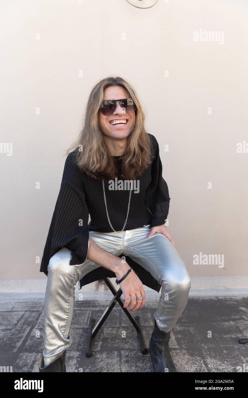 Lachender argentinischer Mann mit langen Haaren, silberne Hose mit Stiefeln und sitzt an einer Wand Stockfoto