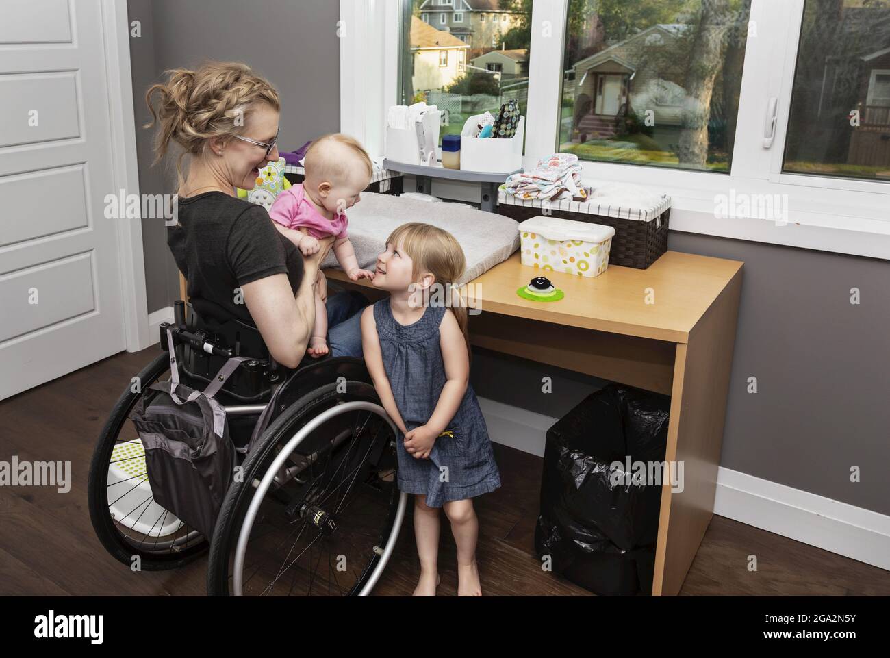 Eine Mutter im Rollstuhl wechselt die Windel ihres Babys, während die große Schwester hilft, das Baby zu amüsieren; Edmonton, Alberta, Kanada Stockfoto