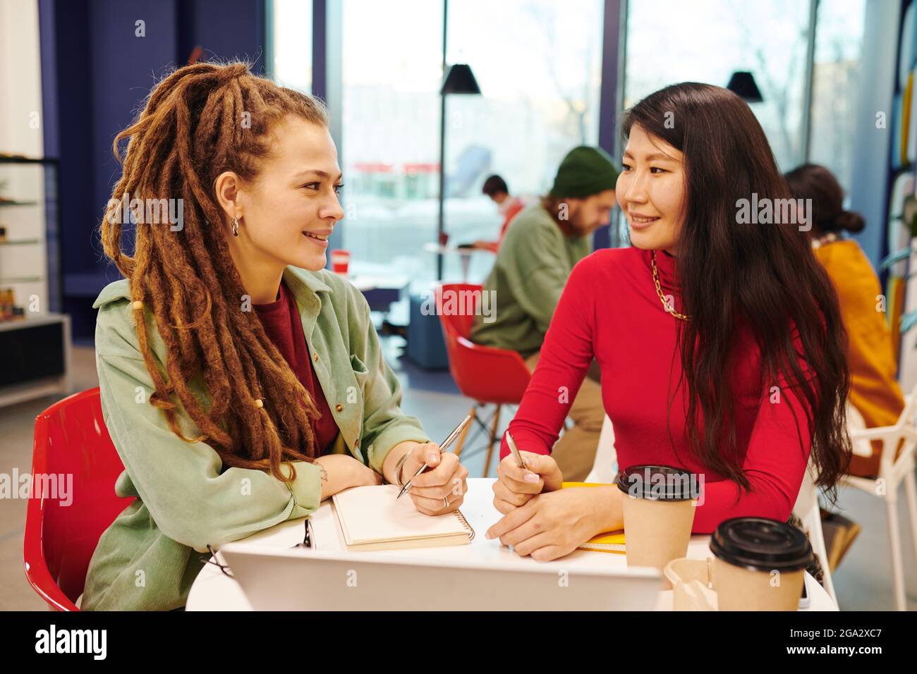 Zwei junge, zeitgenössische interkulturelle Geschäftsfrauen in Casualwear, die über die Präsentation arbeiten Zusammen am Schreibtisch im Büro sitzen Stockfoto