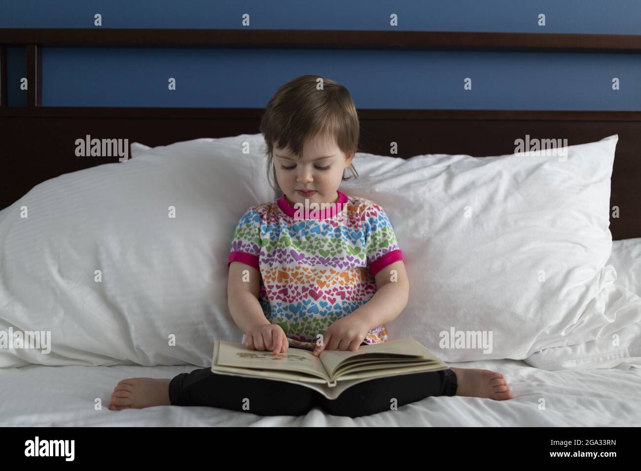 Ein Vorschulmädchen sitzt auf einem Bett und liest ein Buch; White Rock, British Columbia, Kanada Stockfoto