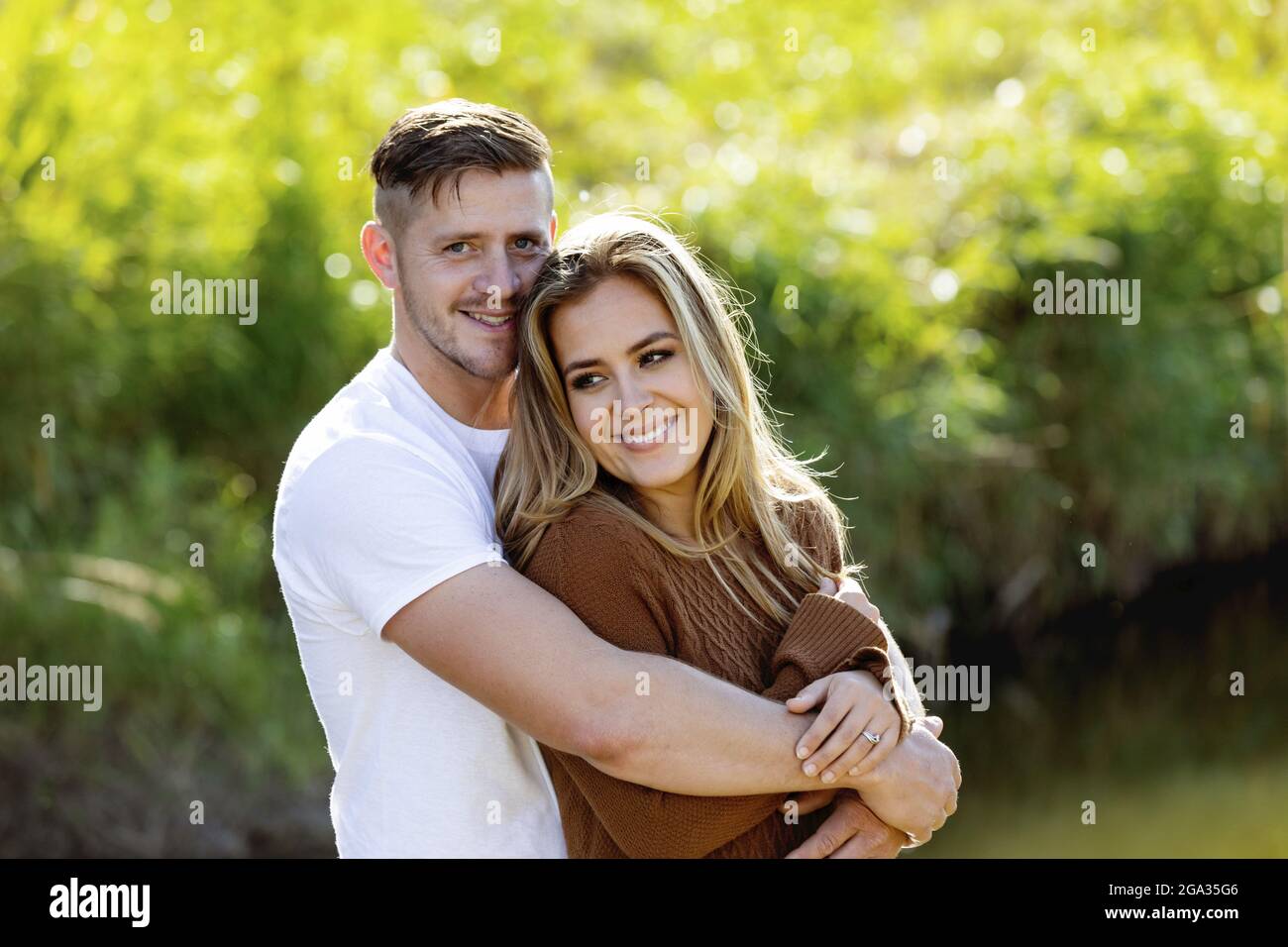Eine kurze Aufnahme eines jungen Ehepaares, das fröhlich lächelt und sich umarmt, während es tagsüber in einem Stadtpark zusammensteht Stockfoto
