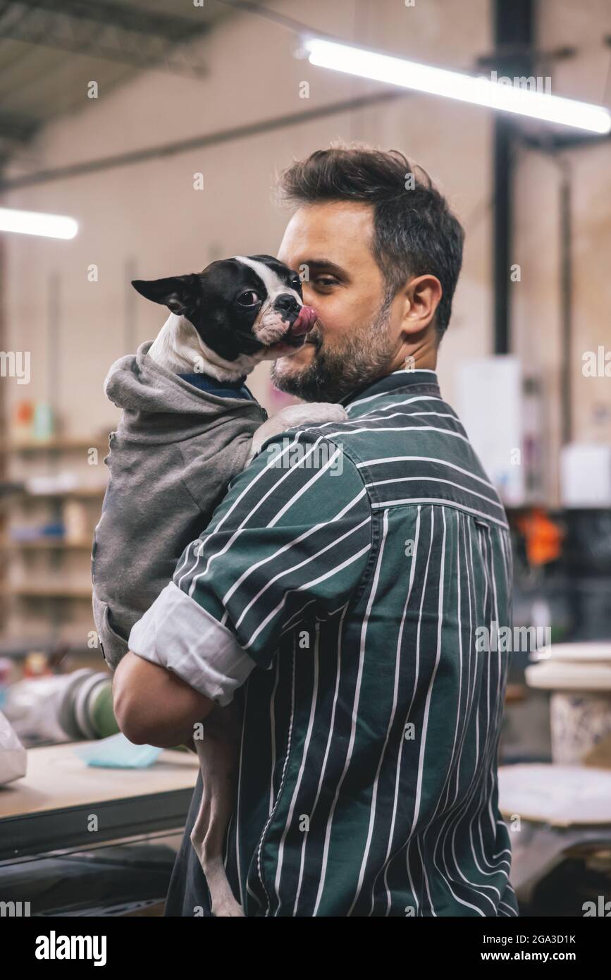 Ein erwachsener hispanischer Mann umarmt eine französische Bulldogge in Kleidung in einer Werkstatt Stockfoto