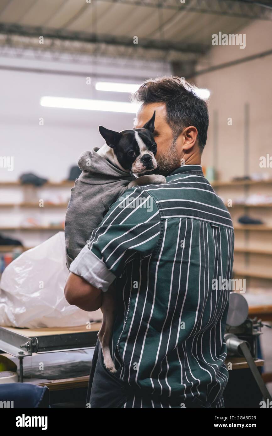 Ein erwachsener hispanischer Mann umarmt eine französische Bulldogge in Kleidung in einer Werkstatt Stockfoto