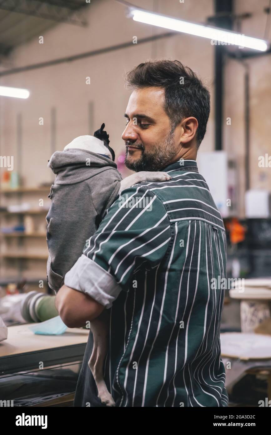 Ein erwachsener hispanischer Mann umarmt eine französische Bulldogge in Kleidung in einer Werkstatt Stockfoto