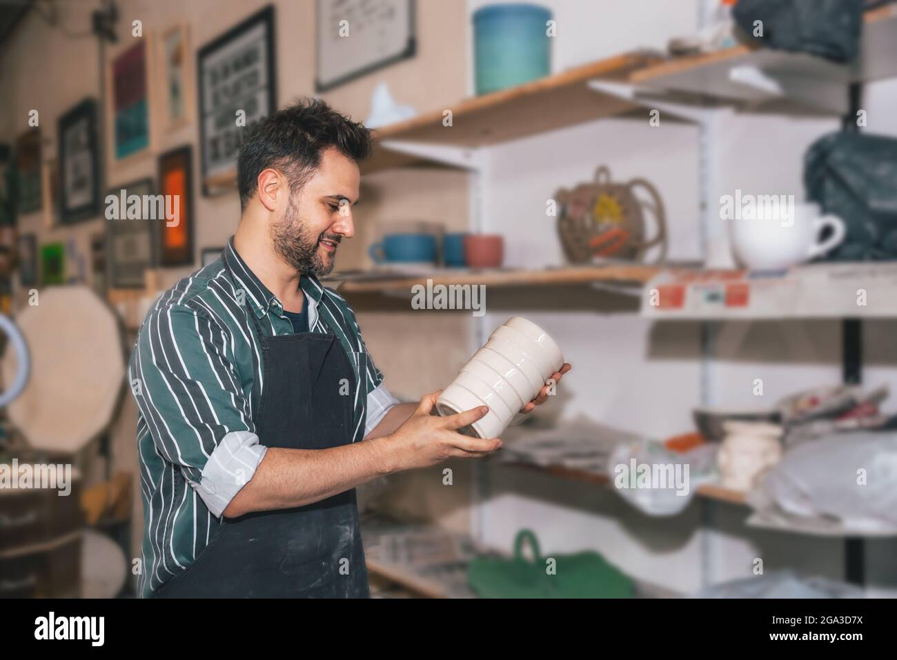 Ein lächelnder Hispanic-Mann mit einer Schürze und einem handgefertigten Tontopf in einer Werkstatt Stockfoto