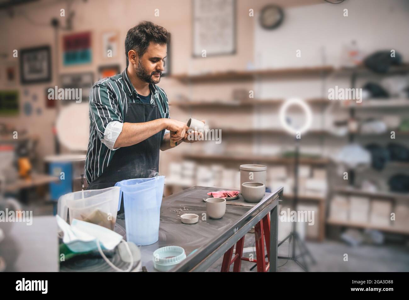 Ein erwachsener hispanischer Mann trägt eine Schürze und schnitzt in einer Werkstatt einen Tontopf über einem Tisch Stockfoto