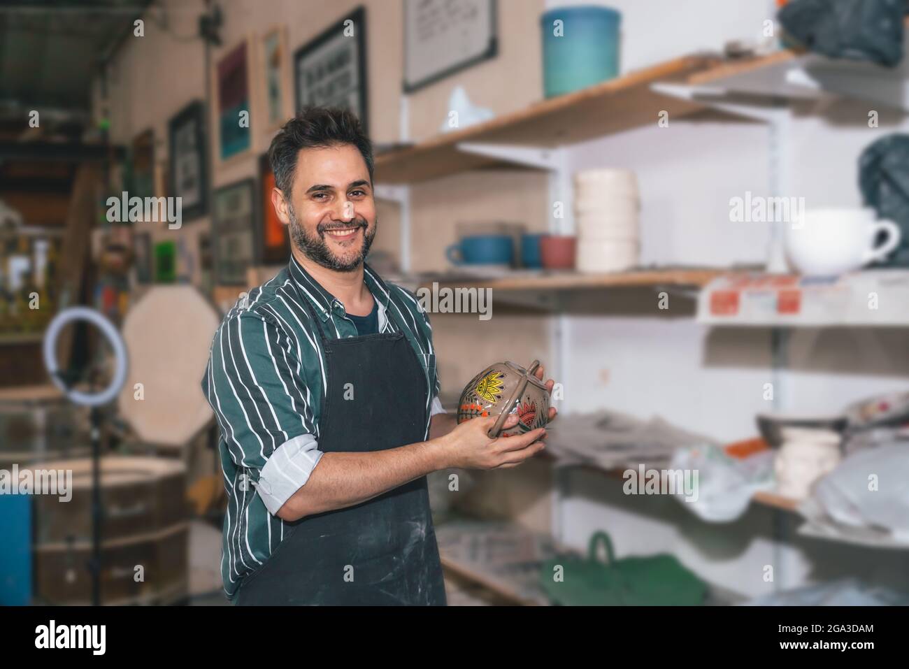 Ein lächelnder Hispanic-Mann mit einer Schürze und einem handgefertigten Tontopf in einer Werkstatt Stockfoto