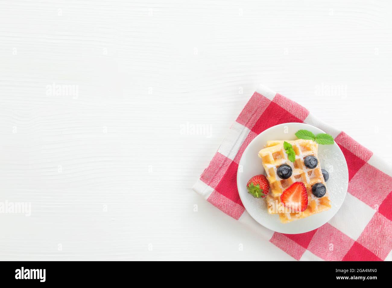 Weißer Teller auf rotem Küchentuch mit hausgemachten belgischen Waffeln, Heidelbeere, Erdbeerschnitt, Minzblatt, Zuckerpulver auf grauem Hintergrund Stockfoto