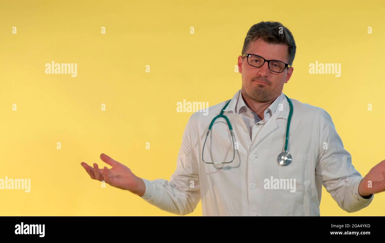 Gutaussehender Arzt im Labormantel, der die Hände auf die Seiten verteilt und zeigt, dass er nichts tut. Gelber Hintergrund. Stockfoto