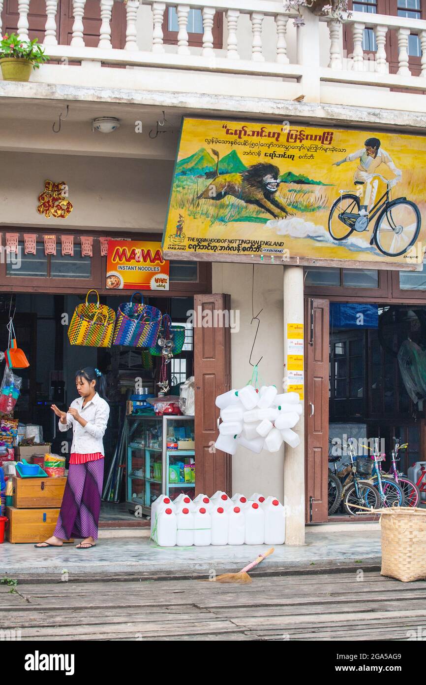 Die junge burmesische Frau mit Lunghi stand vor dem Hardware- und Fahrradladen mit farbenfroher Werbung für den Löwen, der den Mann auf dem Fahrrad jagte, Hsipaw, Myanmar Stockfoto