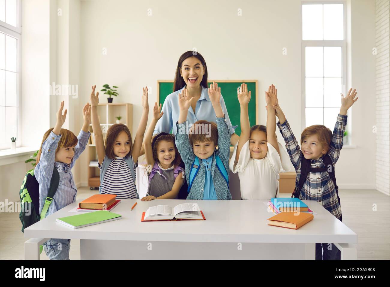Fröhliche Lehrerin und ihre lustigen Grundschüler stehen mit erhobenen Armen. Stockfoto