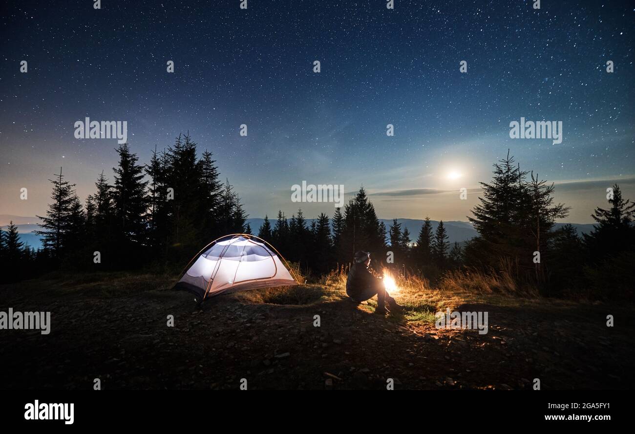 Seitenansicht eines jungen Mannes, der auf Gras in der Nähe seines beleuchteten Zeltes sitzt und die Nachtsterne und den Mond beim Leuchten beobachten kann. Erholung in den Bergen Campingplatz im Freien. Stockfoto