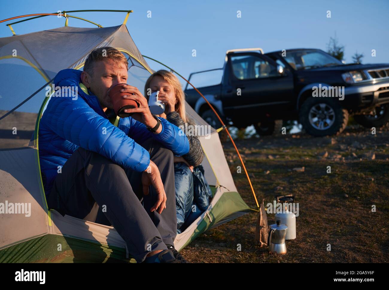 Ein paar Touristen trinken frisch gebrühten Tee sitzen in Zelt auf dem Rasen bei sonnigem Wetter im Frühherbst. Auf dem Hintergrund schwarz SUV geparkt auf Stockfoto