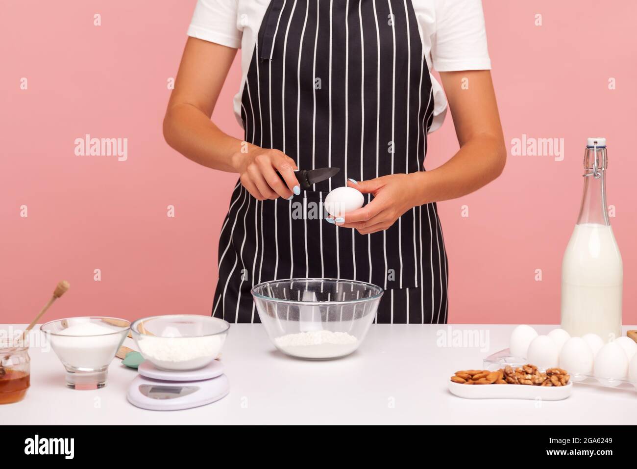 Zugeschnittenes Bild einer gesichtslosen Bäckerin, die Gebäck macht, Eier zum Mehl für die Teigherstellung treibt, schwarze gestreifte Schürze trägt, Zutaten für die Bäckerei. Innen Stockfoto