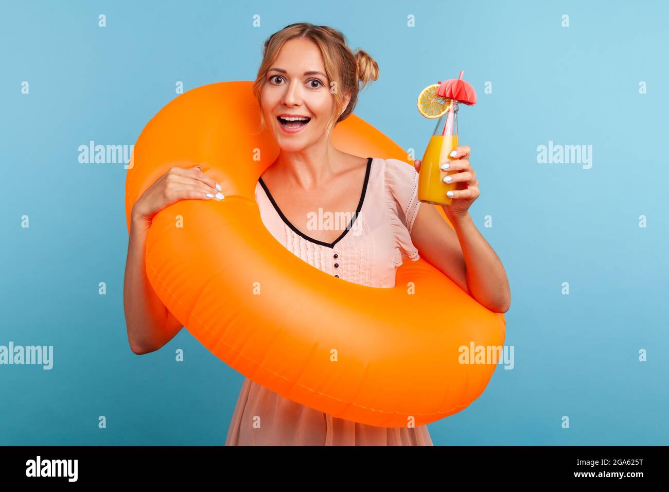 Aufgeregte Frau mit lustigen Haarbrötchen, die sich im Sommerresort entspannen, einen orangenen Gummiring halten und einen frischen Fruchtcocktail genießen und fröhlich über das Vacati lachen Stockfoto