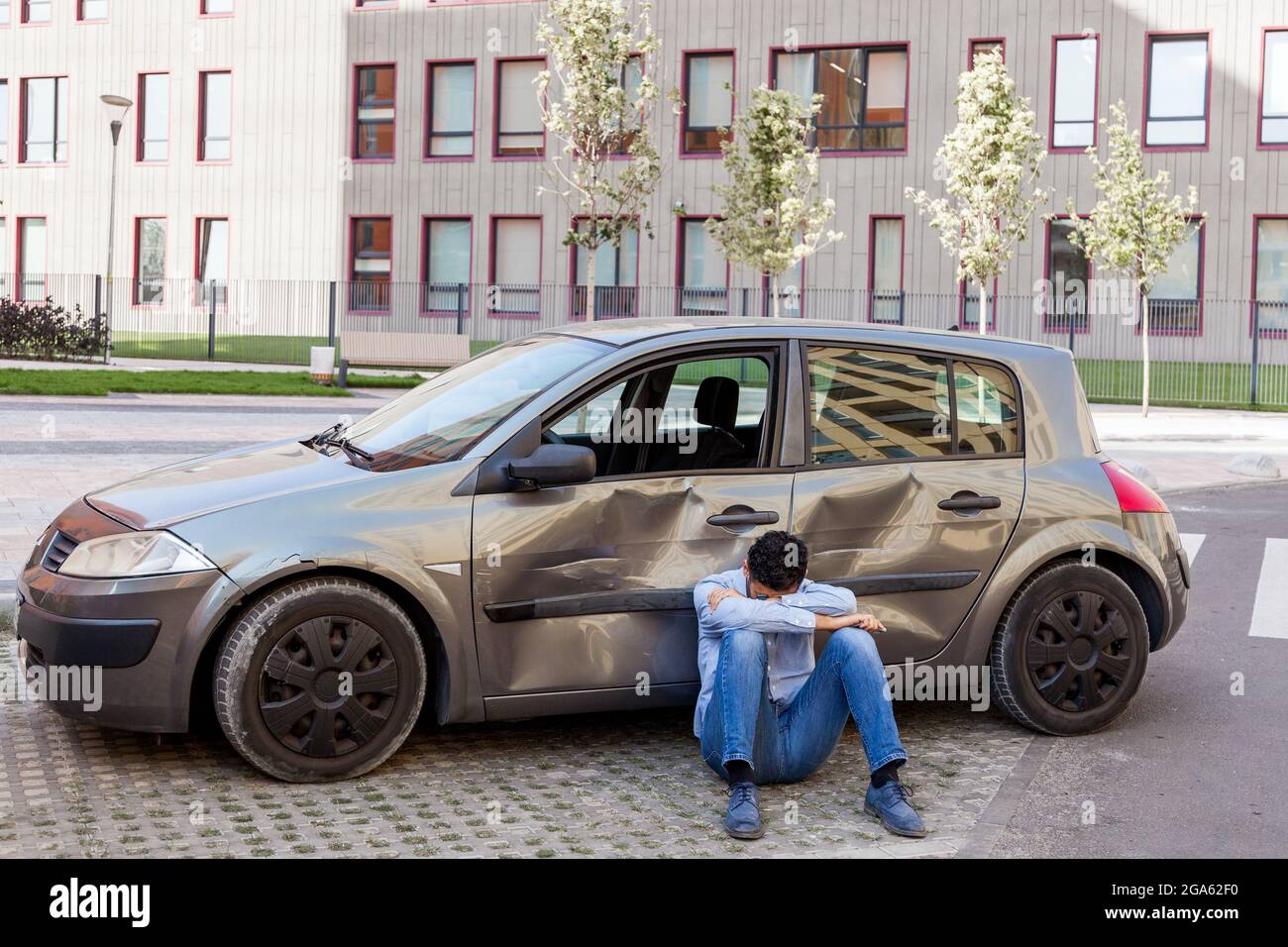 Mann in Verzweiflung trägt Freizeitkleidung auf dem Boden sitzen mit gekipptem Kopf in der Nähe von gebrochenem Auto, beschädigt Auto mit Kratzern und Beulen an Türen, sein Stockfoto