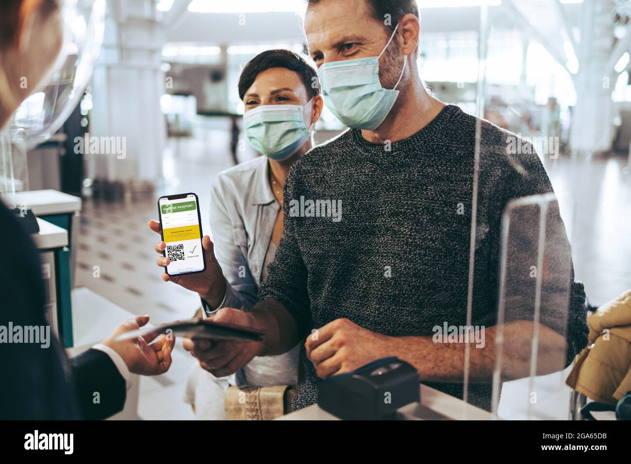 Touristenfamilie, die während einer Pandemie am Check-in-Schalter am Flughafen den Impfpass vorzeigt. Mann, der am Check-in-Schalter steht, während Frau digitale V-Anzeigen vorführt Stockfoto