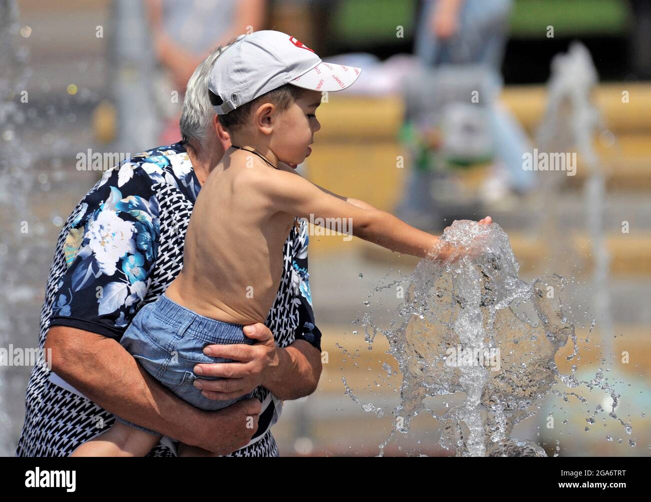 VINNYTSIA, UKRAINE - 28. JULI 2021 - EINE Frau hält einen Jungen, der im Sommer in Vinnytsia, Zentralukraine, einen Wasserstrahl im Sonnensystem-Brunnen berührt. Stockfoto