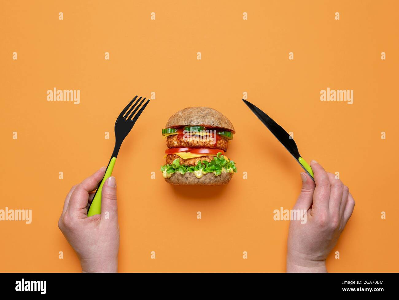 Frau, die veganen Burger isst, Blick von oben auf einen farbigen Tisch. Frau hält Messer und Gabel in Händen und bereitet sich auf den Verzehr eines veganen Sandwiches vor. Stockfoto