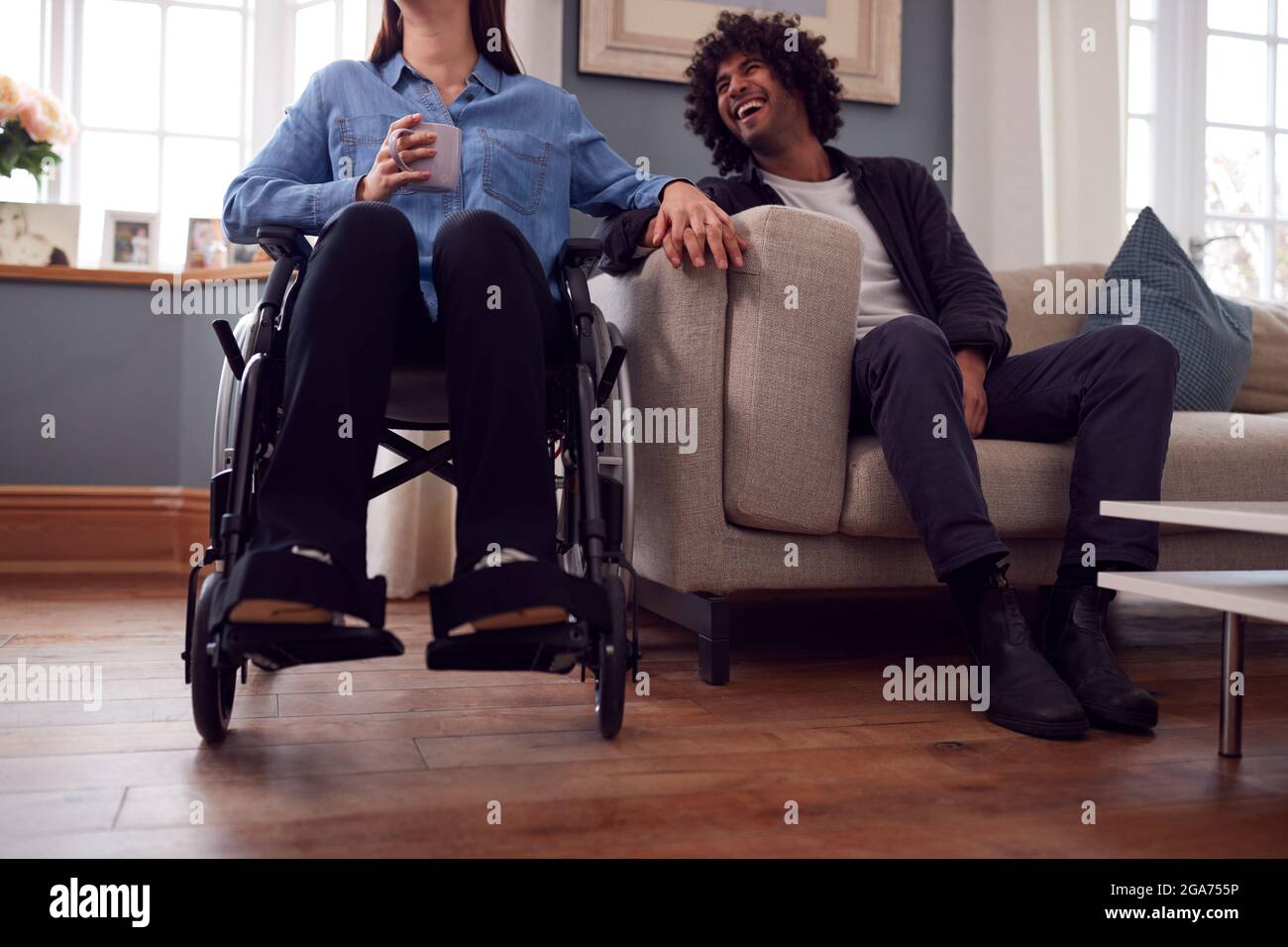 Nahaufnahme Eines Paares Zu Hause Mit Einer Behinderten Frau Im Rollstuhl, Die Hände Hält, Während Der Mann Auf Dem Sofa Sitzt Stockfoto