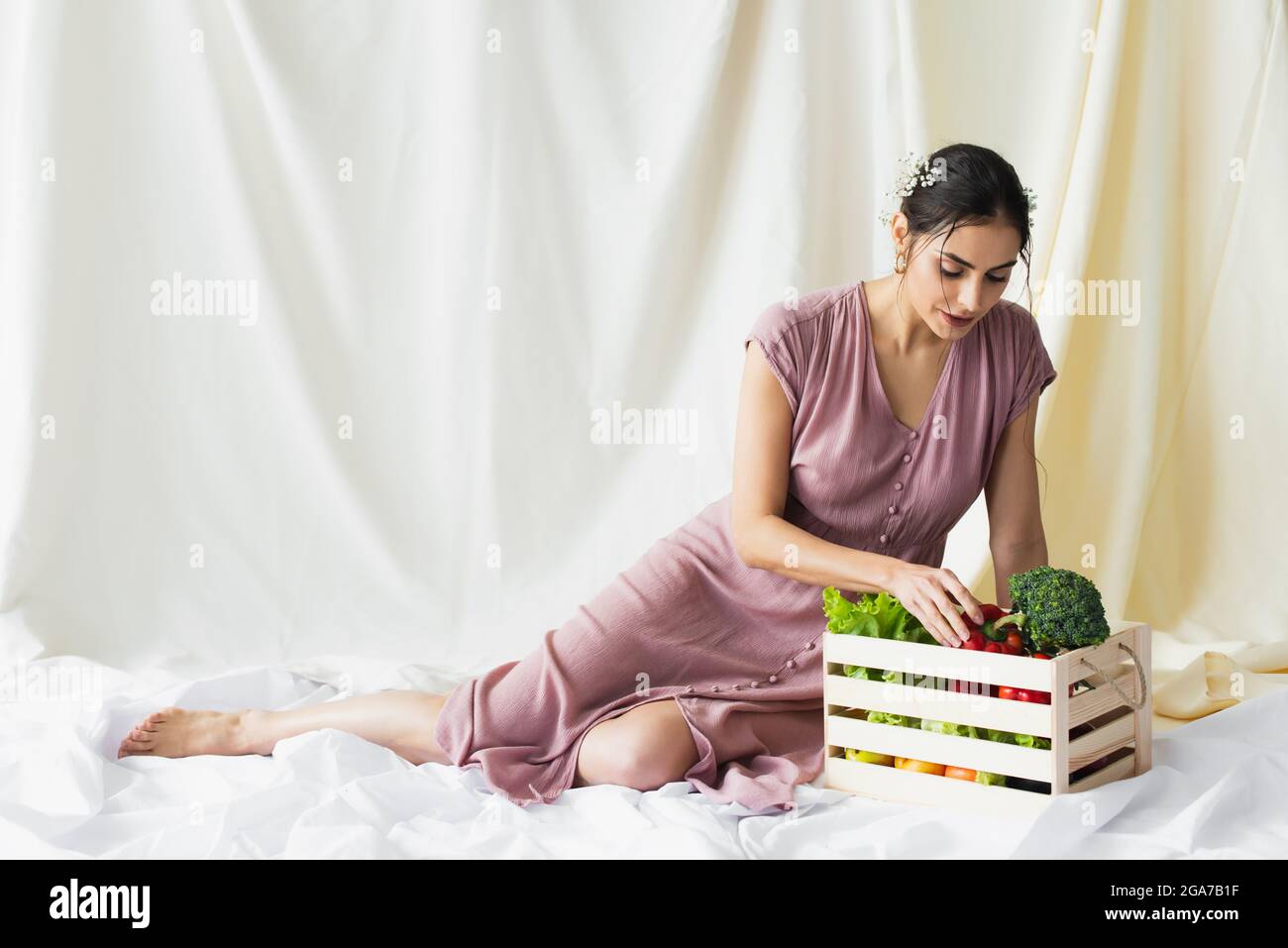 Brunette Frau erreicht roten Paprika in der Nähe von Gemüse in Holzbehälter auf weiß Stockfoto