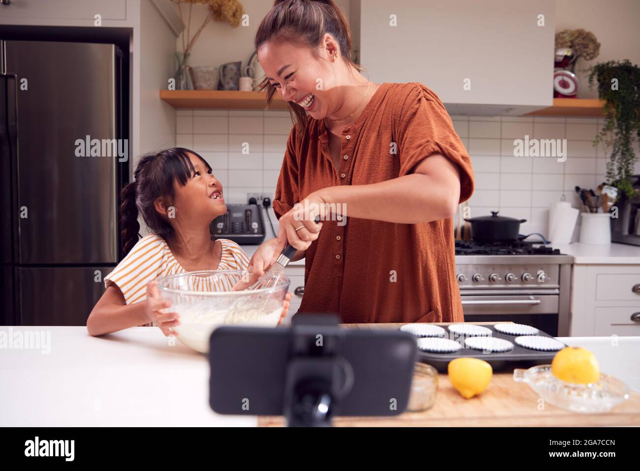 Asiatische Mutter Und Tochter Backen Cupcakes In Der Küche Zu Hause, Während Auf Vlogging Auf Handy Stockfoto