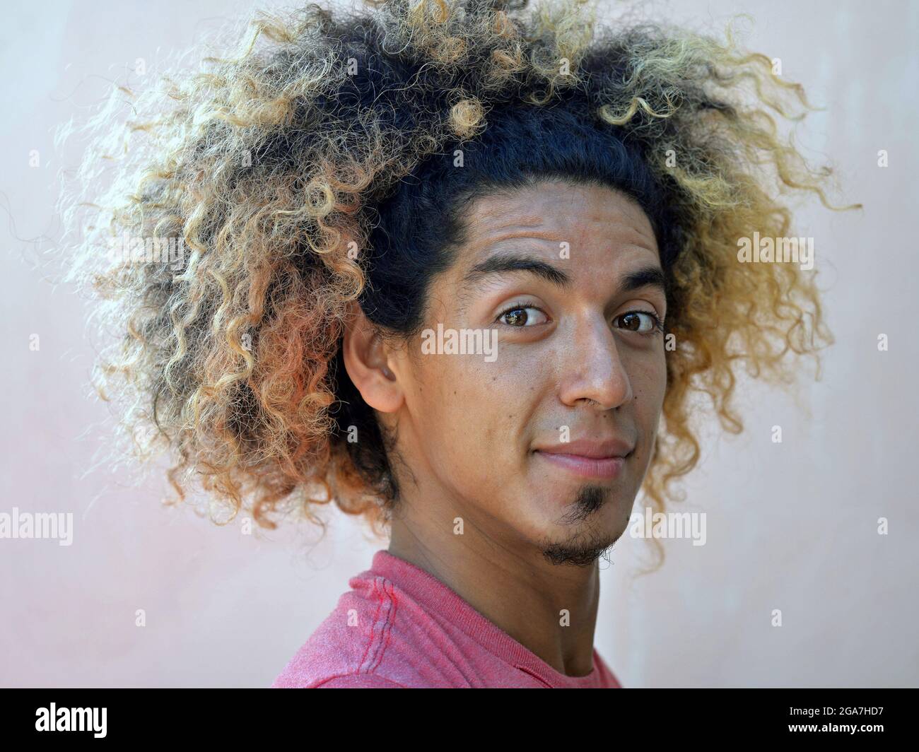 Der junge lateinamerikanische Mann mit einem Schock wilder lockiger Haare und blonden gefärbten Haaren lächelt mit geschlossenem Mund an der Kamera. Stockfoto