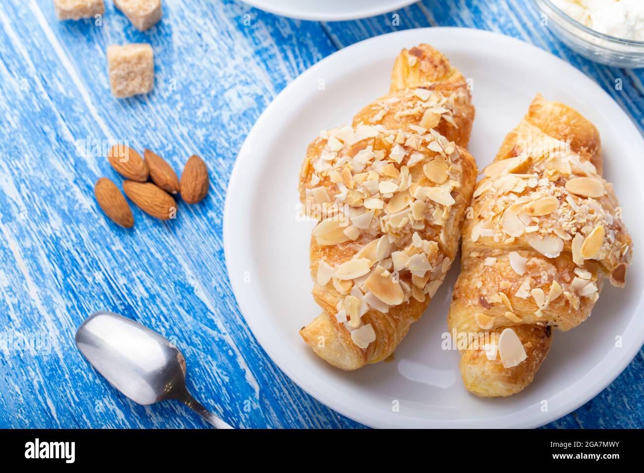 Frühstück am Morgen. Frisches französisches Croissant auf einem Teller. In der Nähe befinden sich verstreute Mandeln und Rohrzucker-Würfel auf einer blauen Holzoberfläche. Traditionelle französische Küche Stockfoto