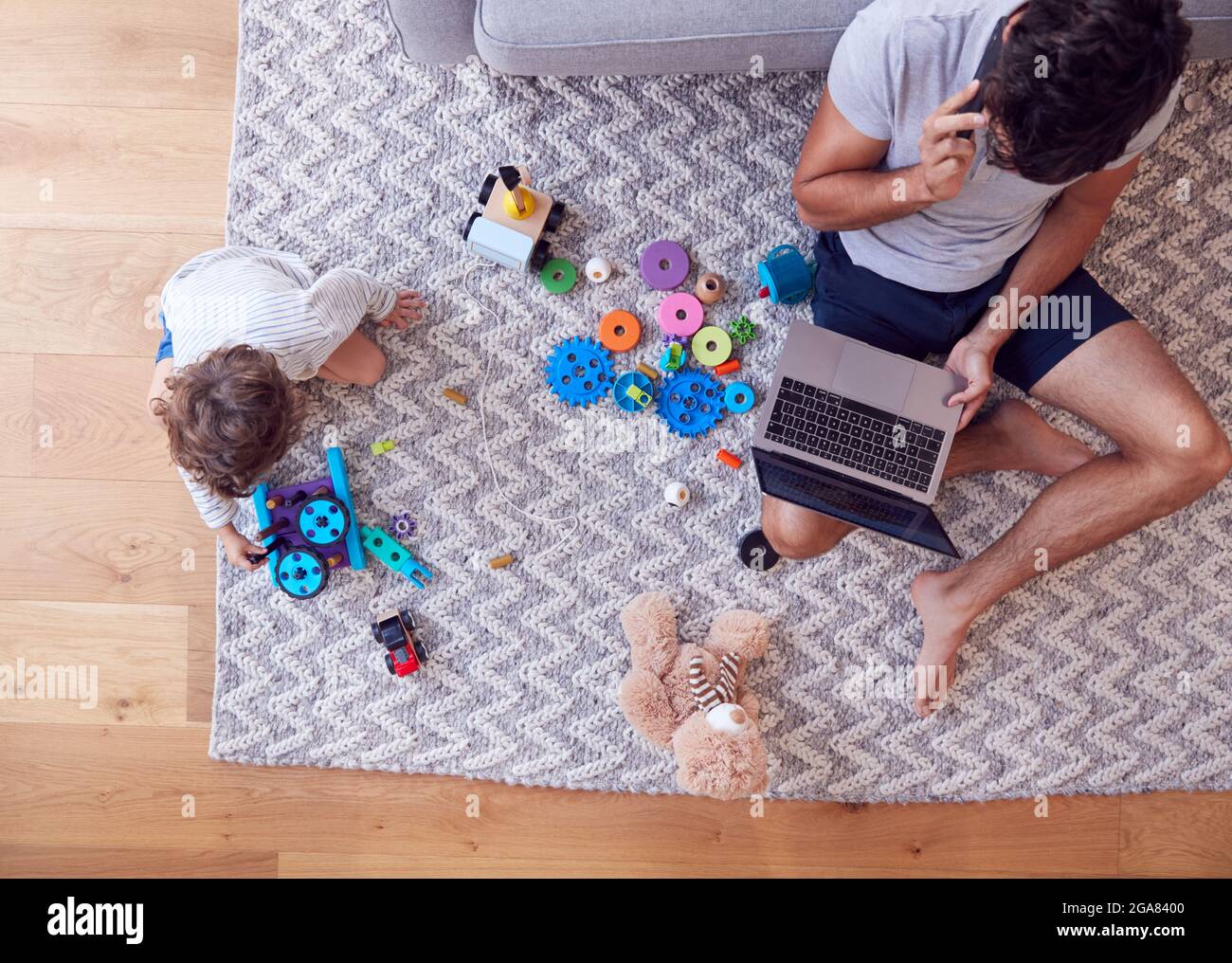 Overhead Shot Of Son Spielt Mit Spielzeug Auf Teppich Zu Hause, Während Vater Nutzt Laptop Und Handy Stockfoto