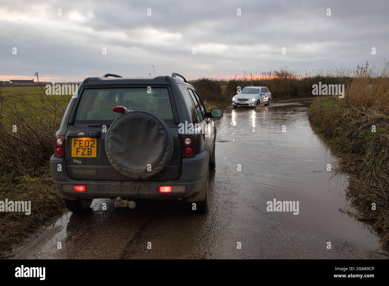 Eine überflutete Straße auf dem Land bei Braunton in North Devon, England, mit einem Auto, das gerade durchfährt, plus einem Land Rover im Vordergrund Stockfoto