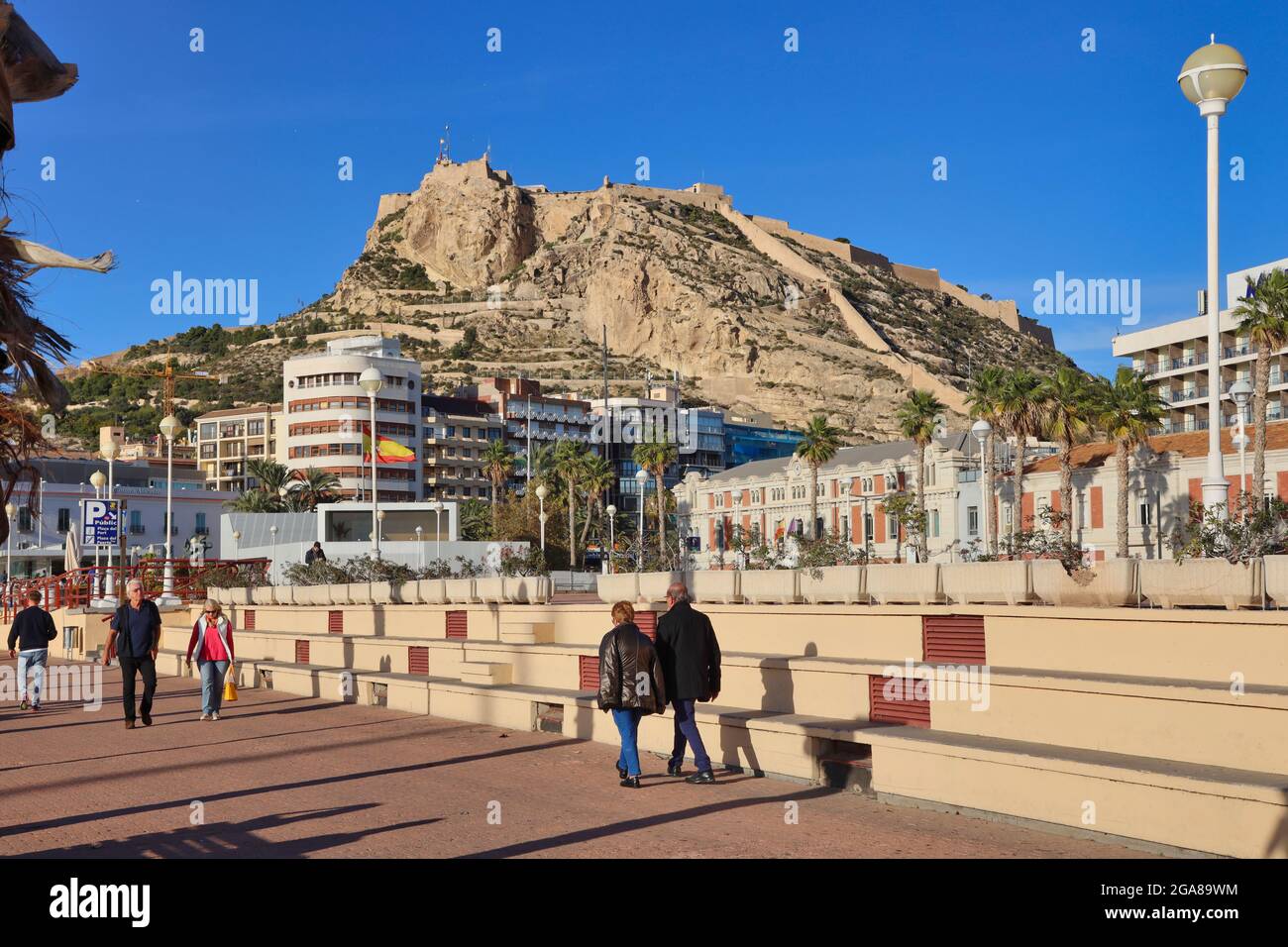 Die Burg von Santa Barbara hoch oben auf einem Vorgebirge mit Blick auf die Stadt Alicante, Spanien, mit der Promenade und den Menschen, die im Vordergrund gehen Stockfoto